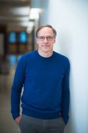 A man in a blue sweater stands against a white wall.